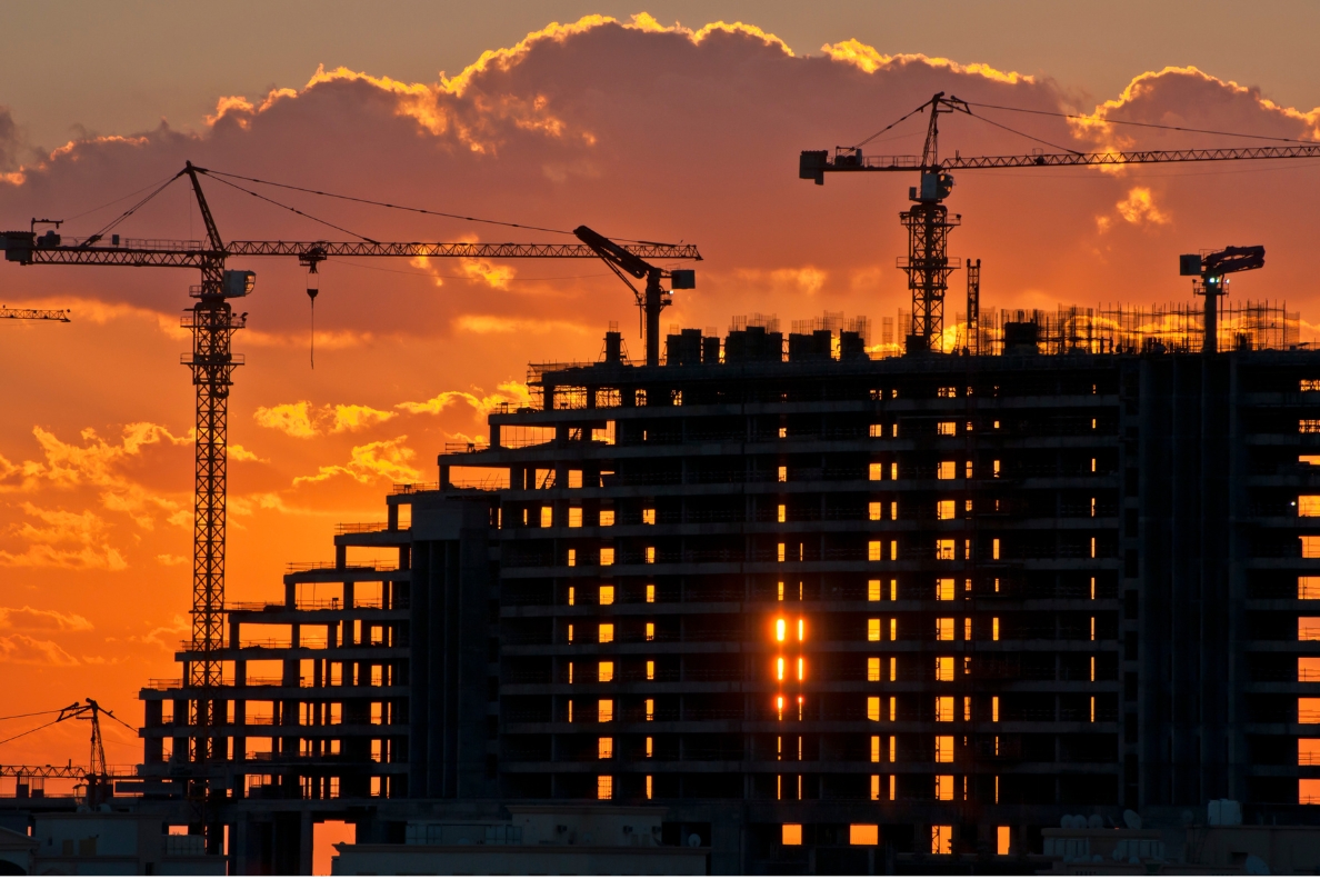 Image of a building site of a high multi-storey building at sunset, with a lot of cranes scattered around.